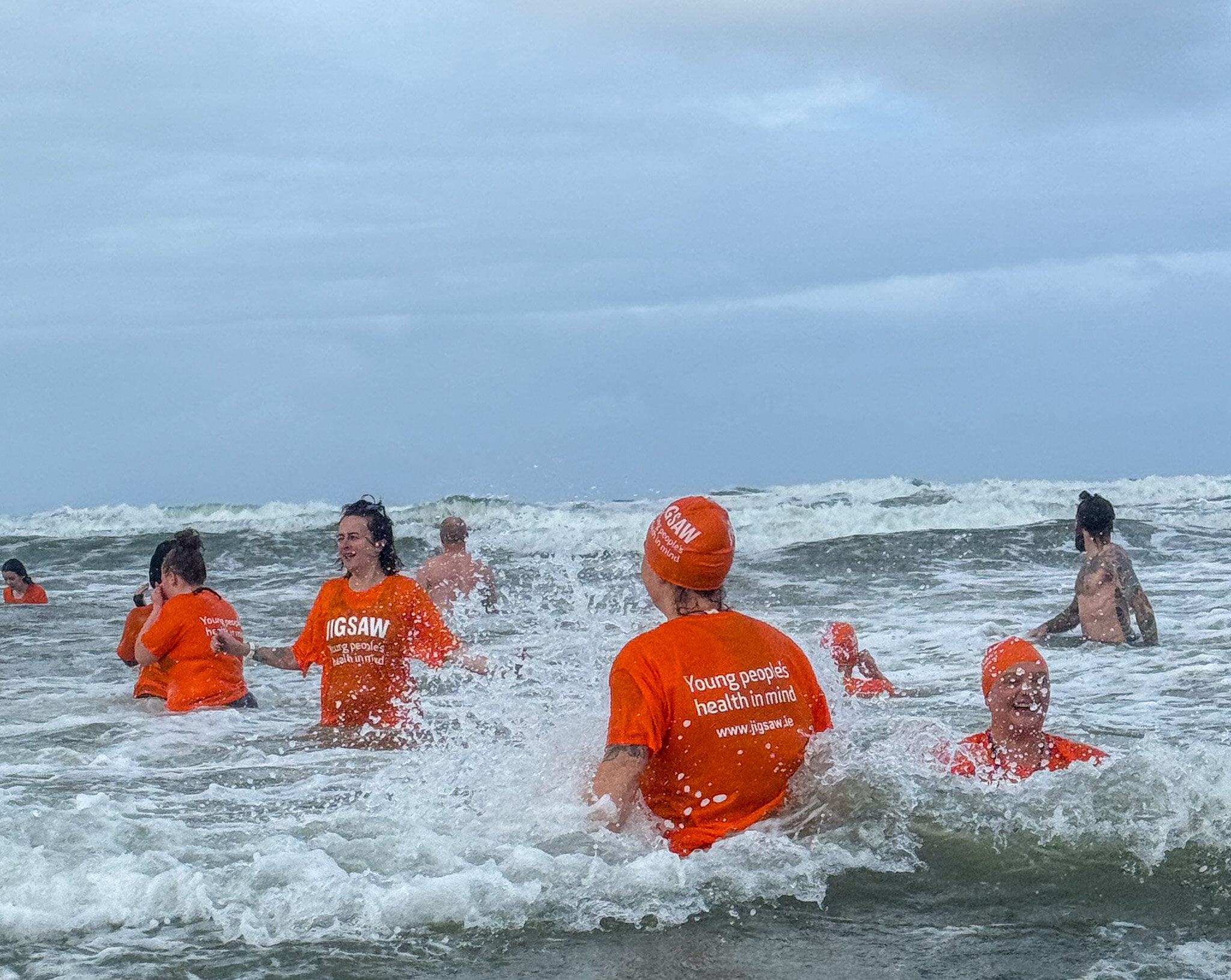 Swimmers in orange Jigsaw t-shirts and swim caps laughing in the waves at Rossnowlagh Beach